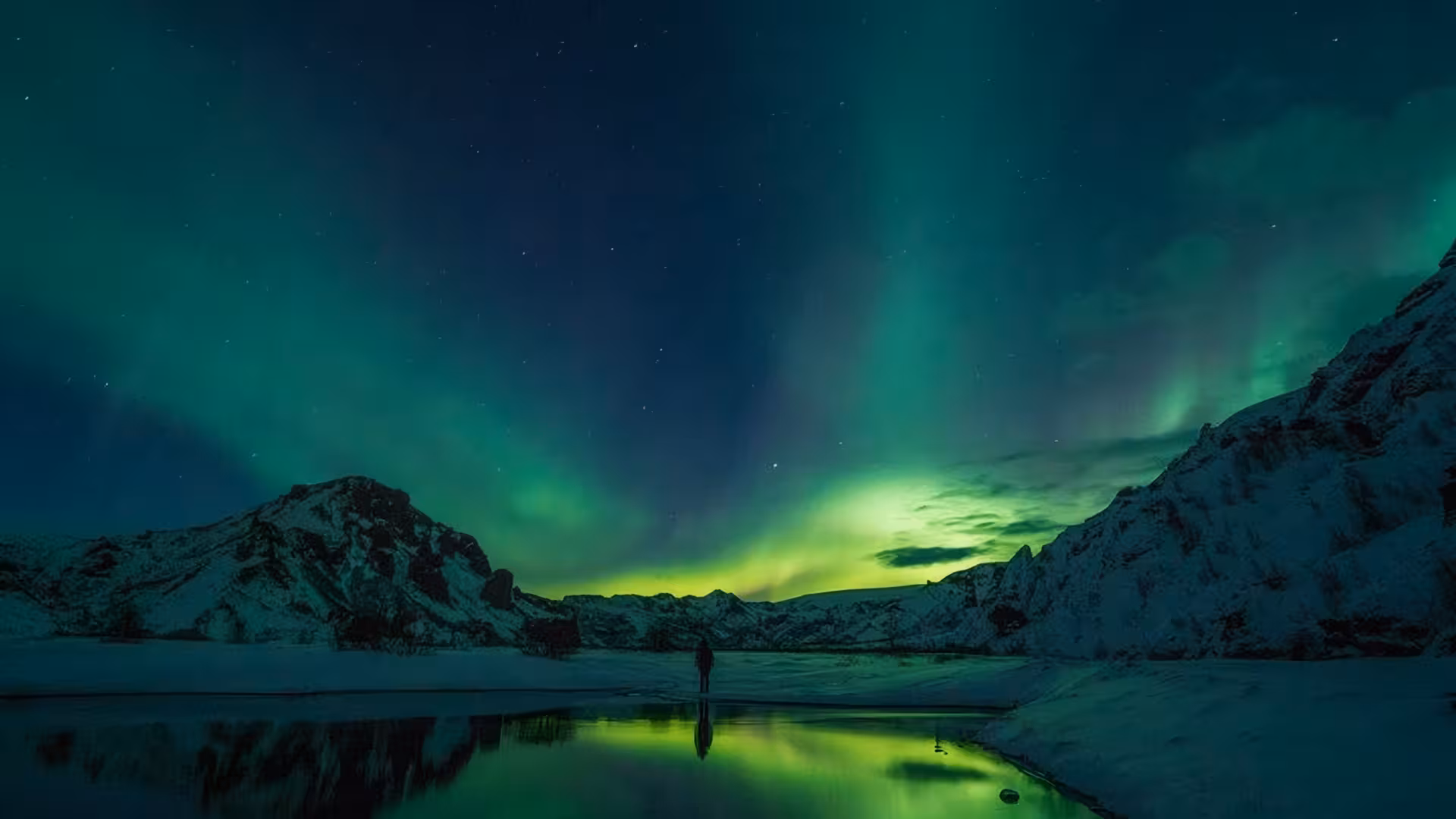 Person standing under vibrant green northern lights aurora borealis over snowy Iceland mountains and reflective lake