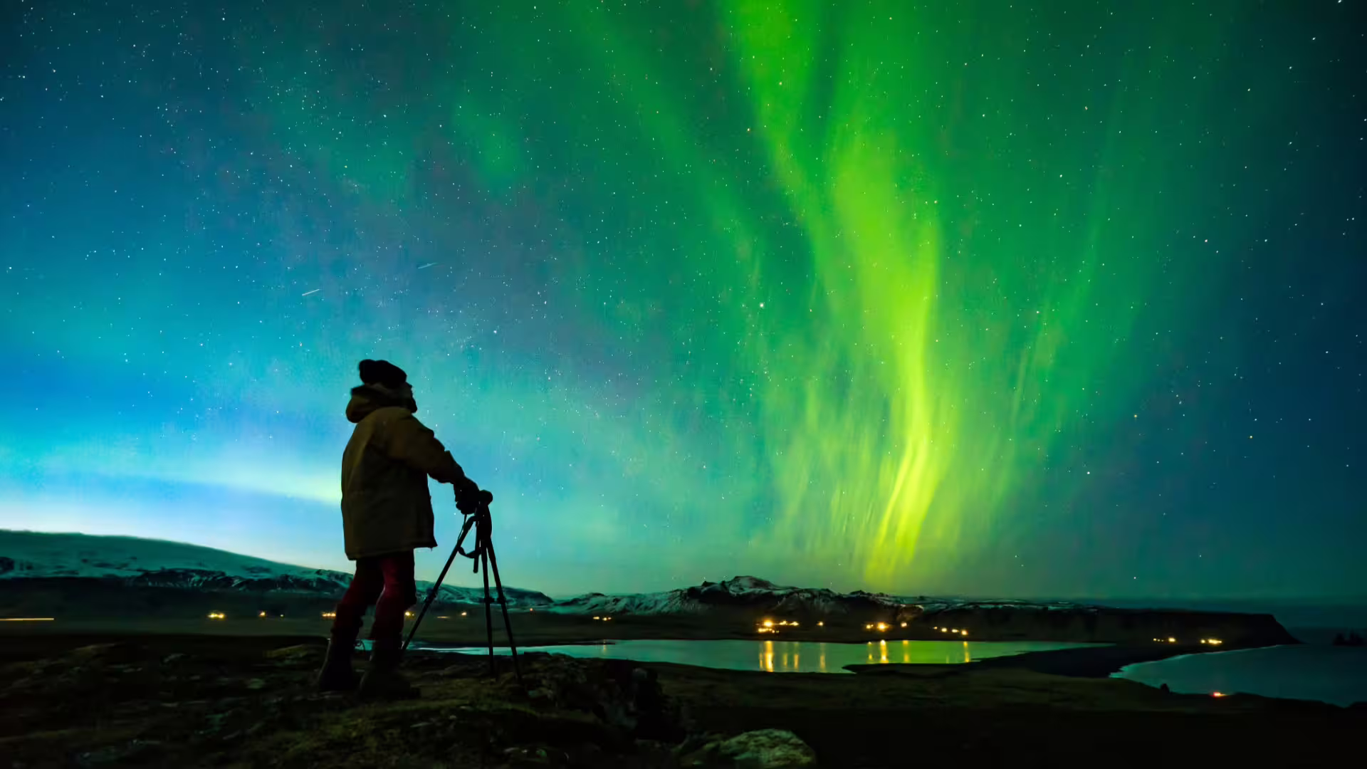 Photographer with tripod standing under vivid green northern lights over Iceland's snowy landscape at night