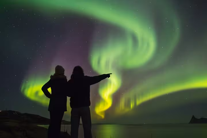 Two silhouettes watching vibrant green and purple Northern Lights swirling over Reykjavik waterfront at night