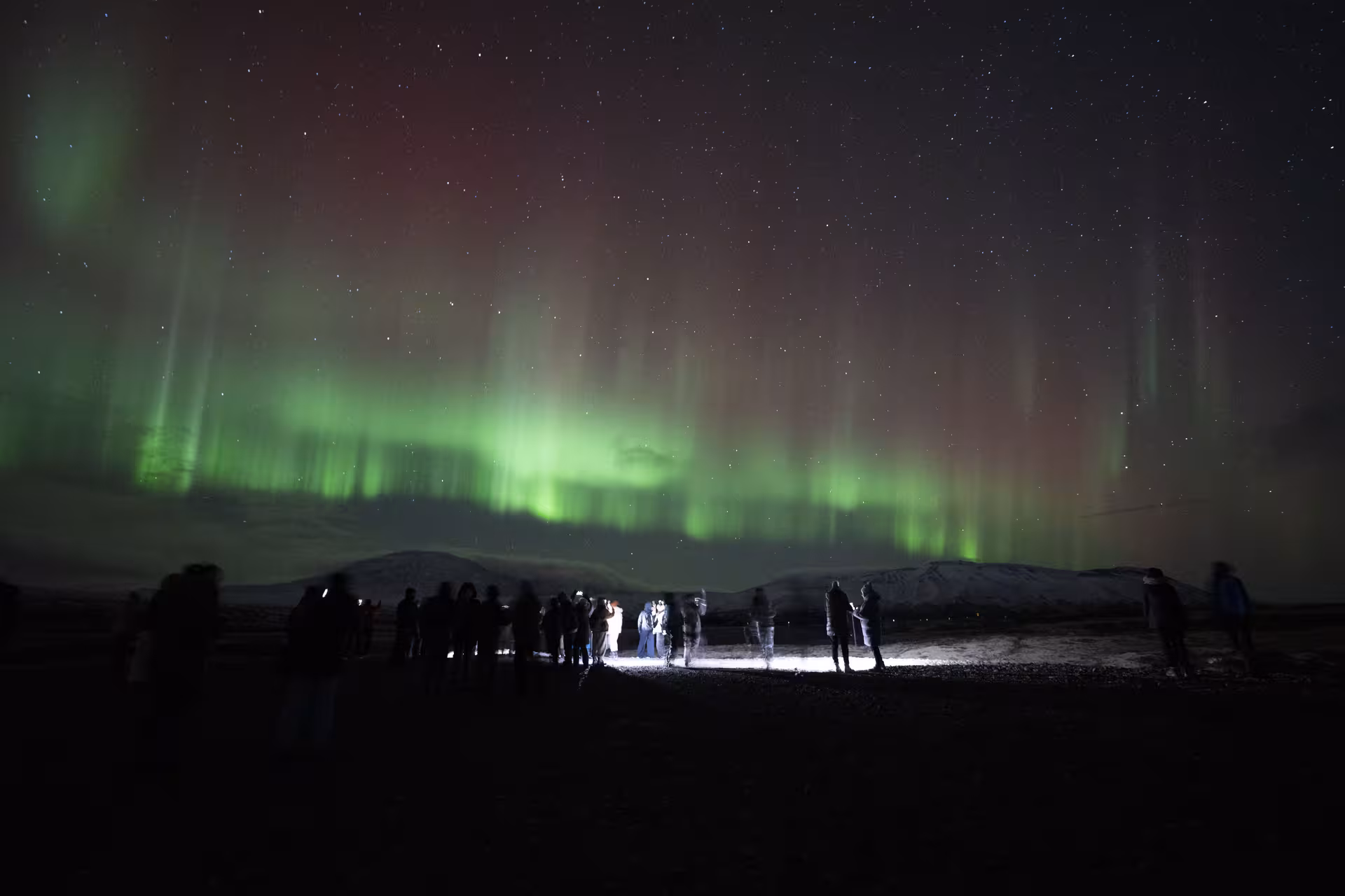 Group of tourists watching vibrant green and red northern lights over snowy Iceland landscape at night