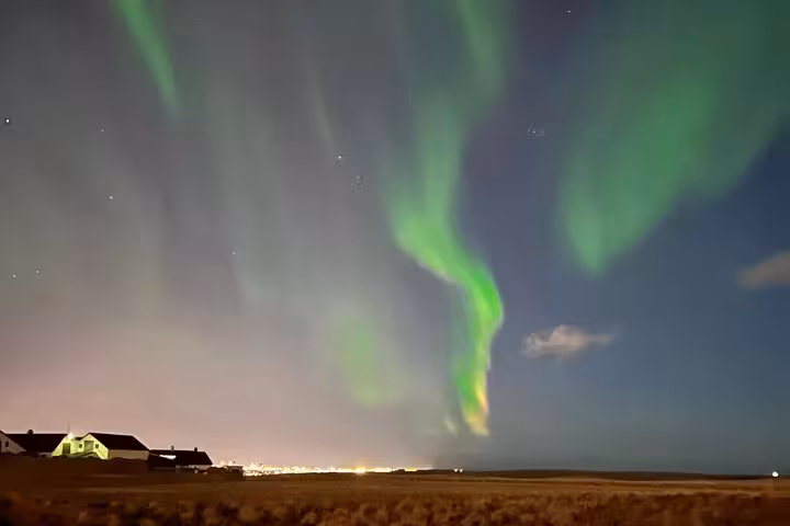 Vibrant green northern lights dancing over flat Icelandic landscape with illuminated buildings at night