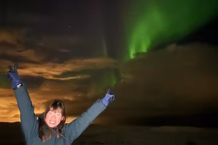 Excited woman with arms raised pointing at vibrant green northern lights aurora borealis over cloudy Reykjavik night sky
