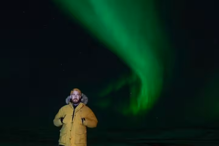 Man in yellow jacket standing under vibrant green northern lights in Reykjavik night sky
