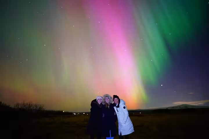 Three people posing under vibrant green and pink Northern Lights aurora borealis in Reykjavik Iceland