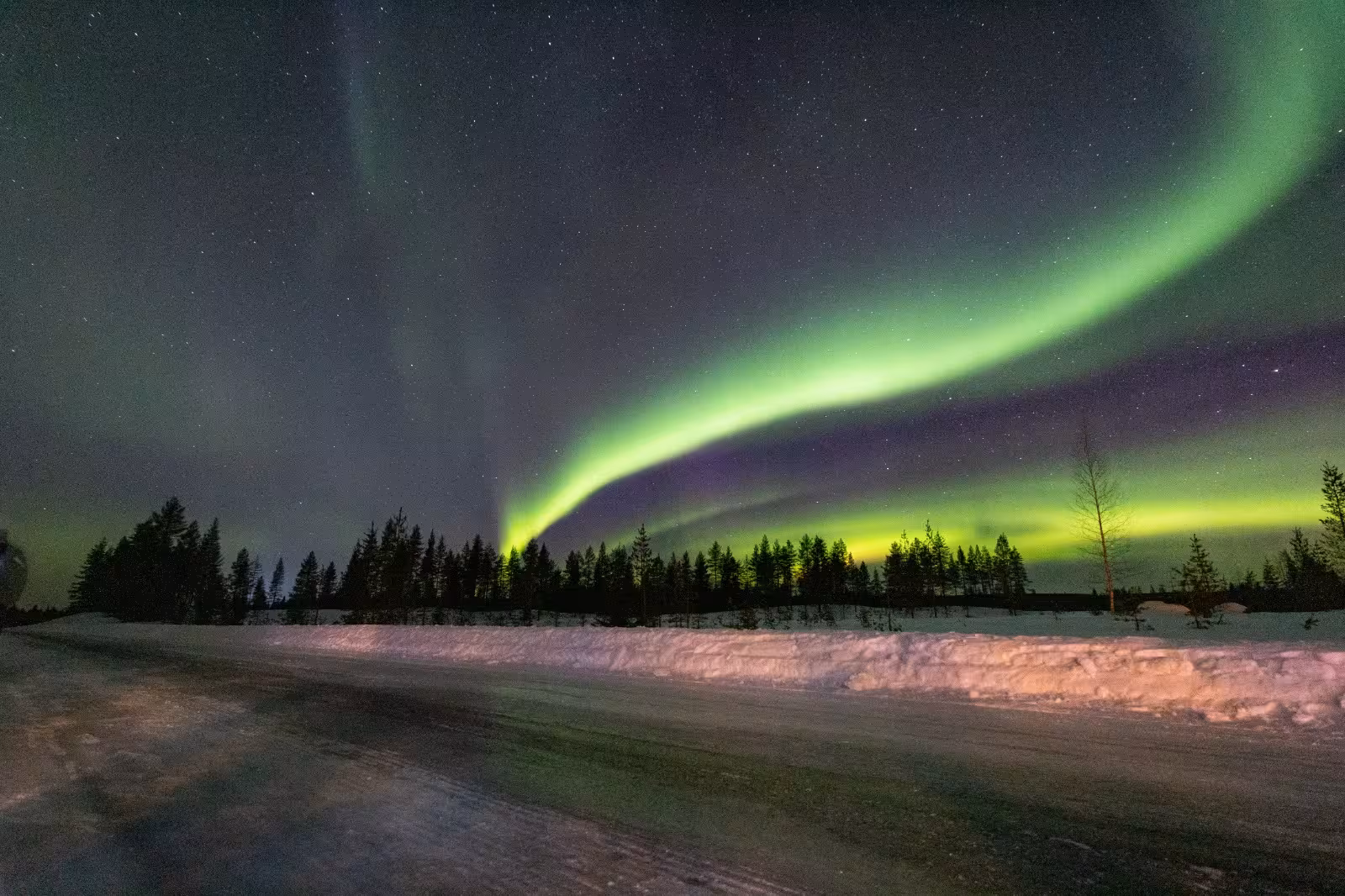 Stunning aurora borealis illuminating a winter night sky above a serene, snow-covered road and forest.