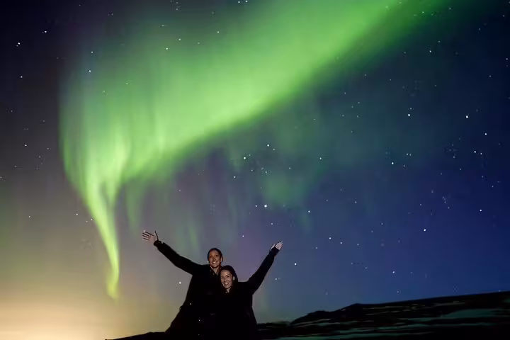 Two excited travelers posing under vibrant green northern lights aurora borealis in Iceland night sky