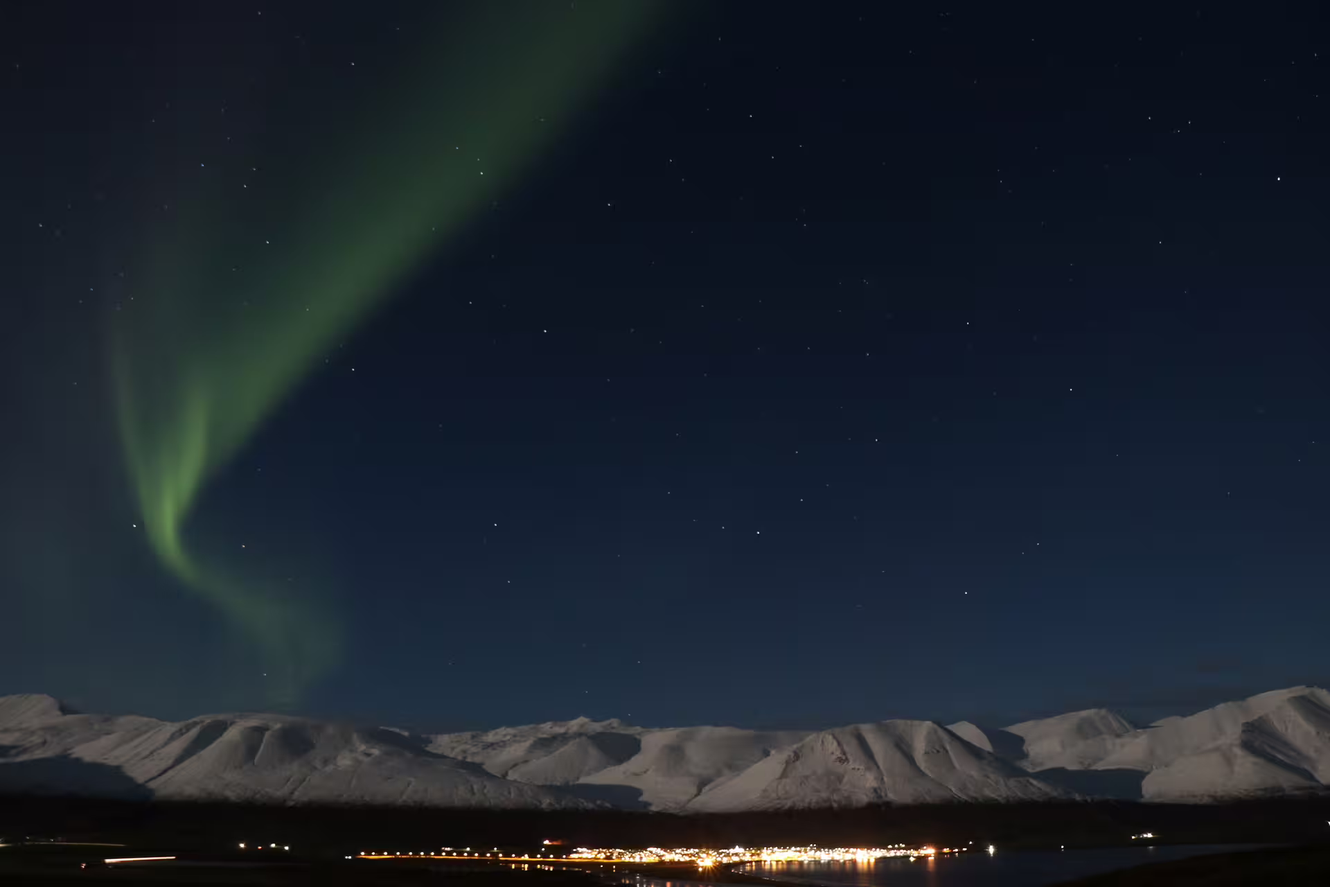 Northern lights dancing over snow-covered mountains above a lit Icelandic town at night