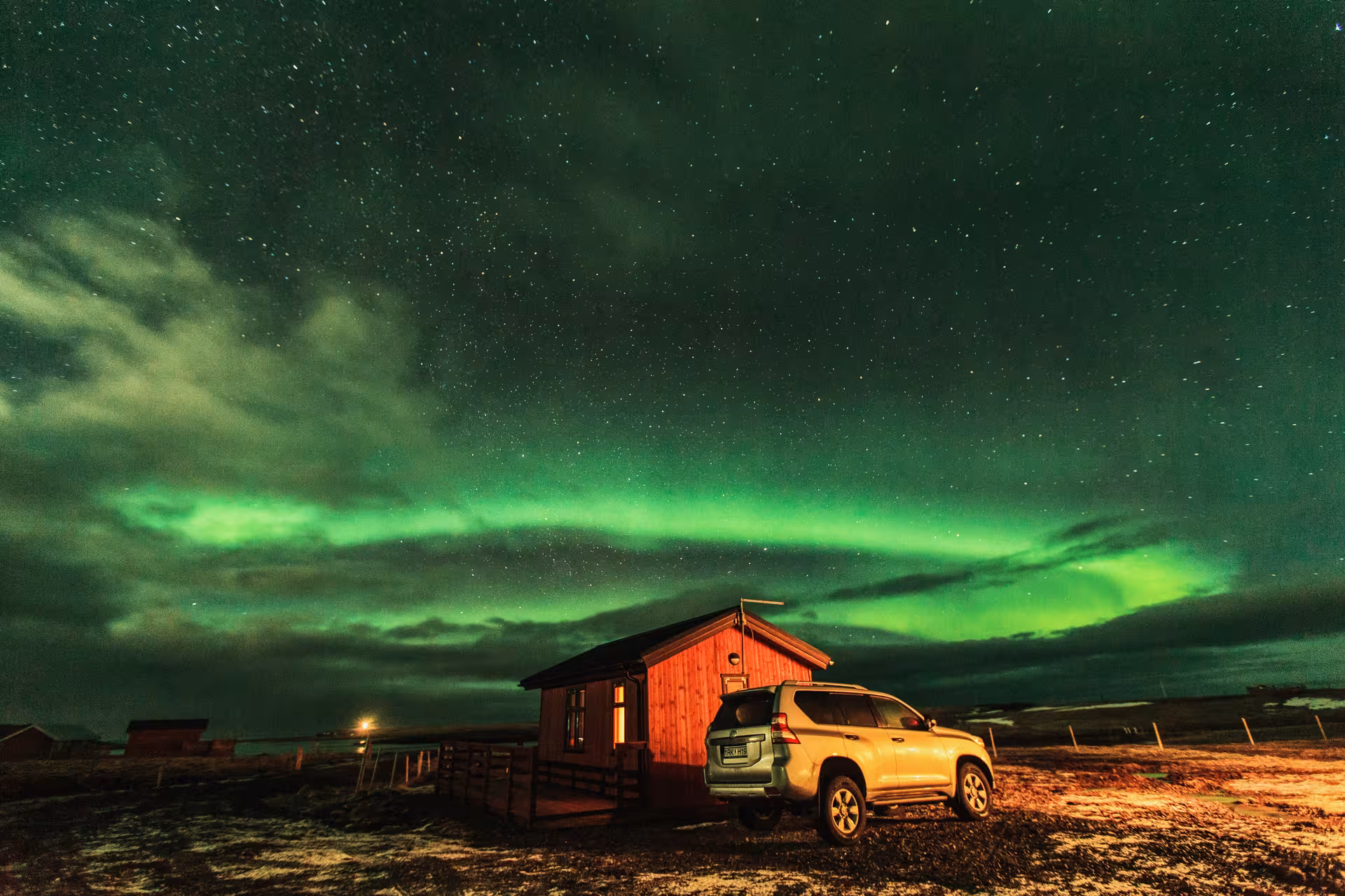 Aurora borealis glowing green over a red cabin with a luxury SUV in Iceland at night