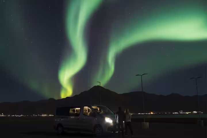 Two people standing by a van watching vibrant green northern lights over mountains near Reykjavik