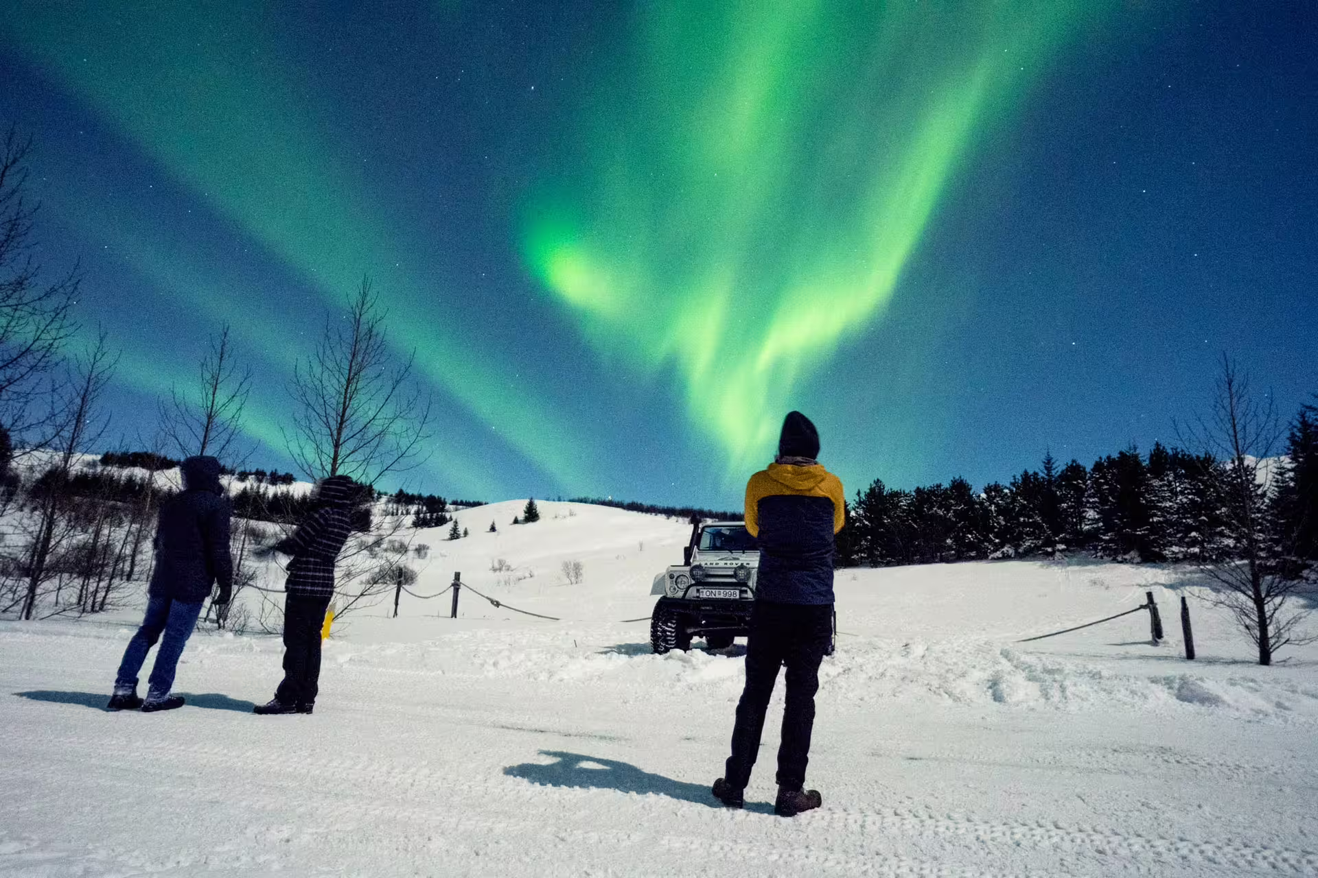 Three people watching vivid green northern lights over snowy Iceland landscape beside a Super Jeep at night