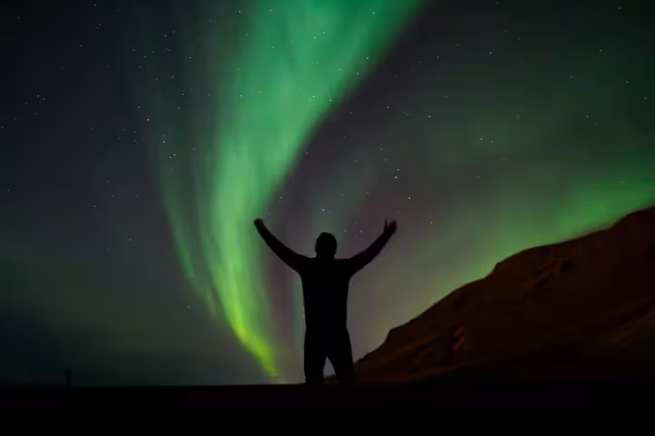 Silhouette of person with arms raised under vibrant green aurora borealis in Iceland night sky