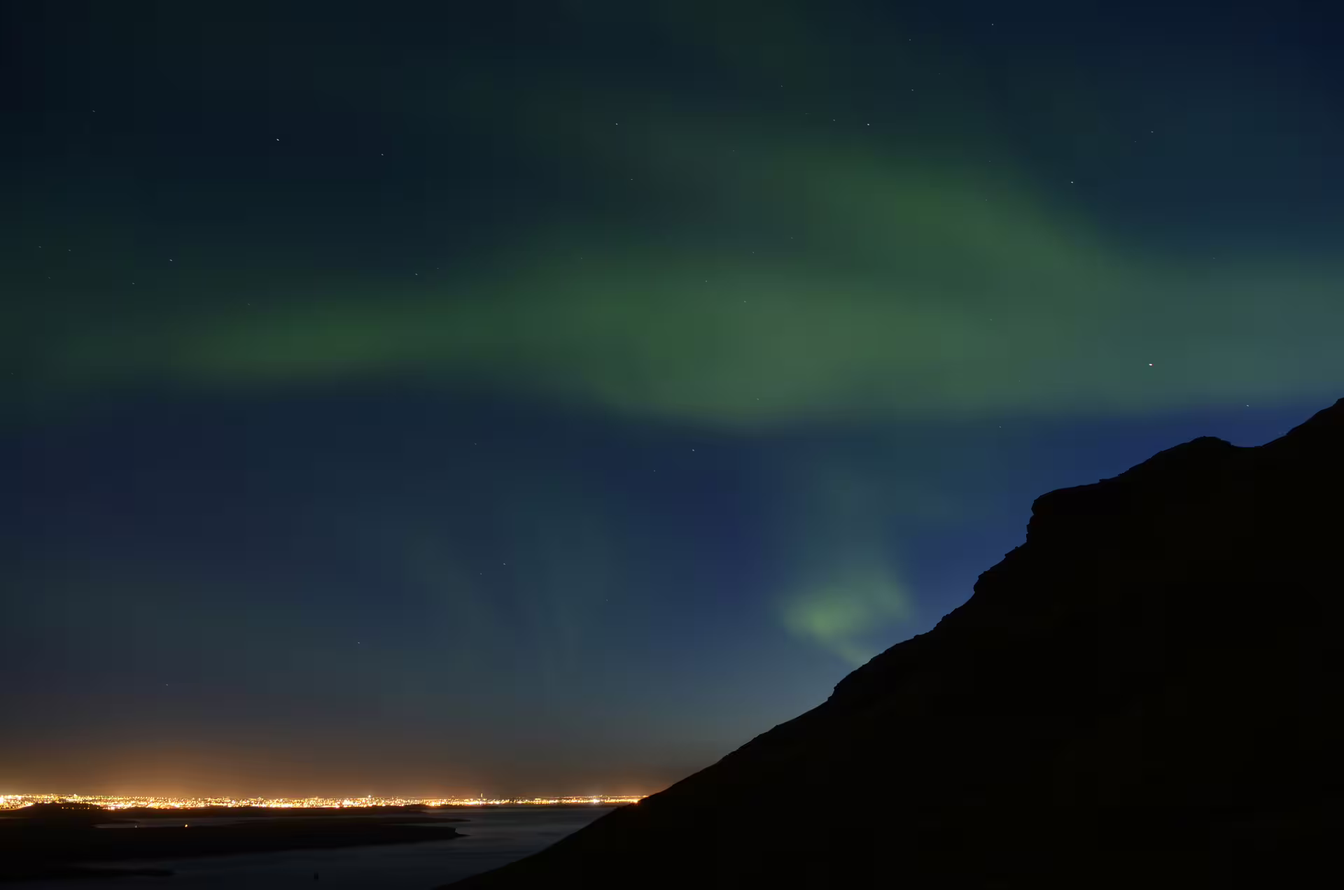 Northern lights glowing green over Reykjavik Bay with city lights visible along the coastline at night