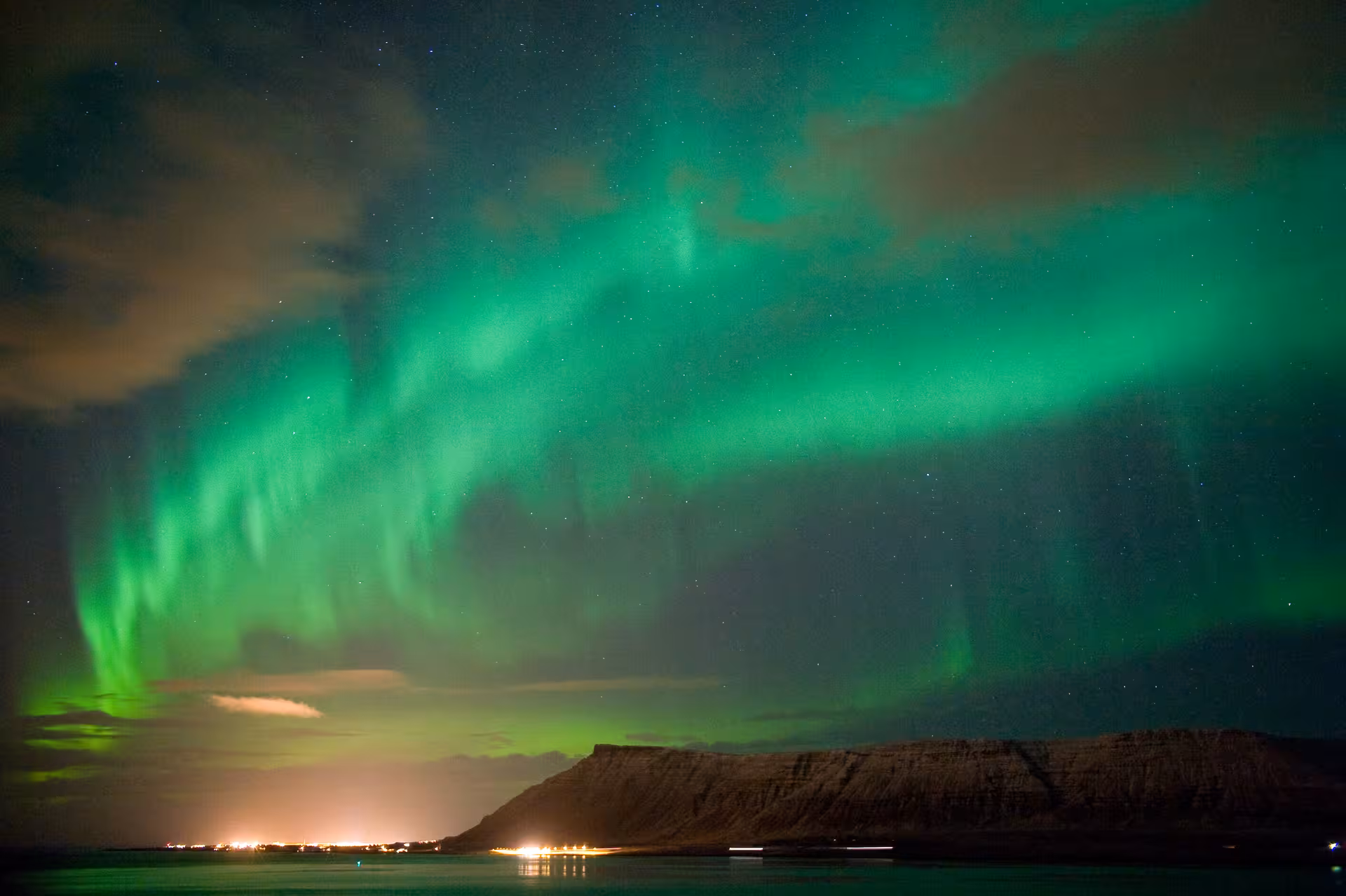 Stunning Northern Lights illuminate the sky over Reykjavík's coastline, with vibrant green hues reflecting on the water.