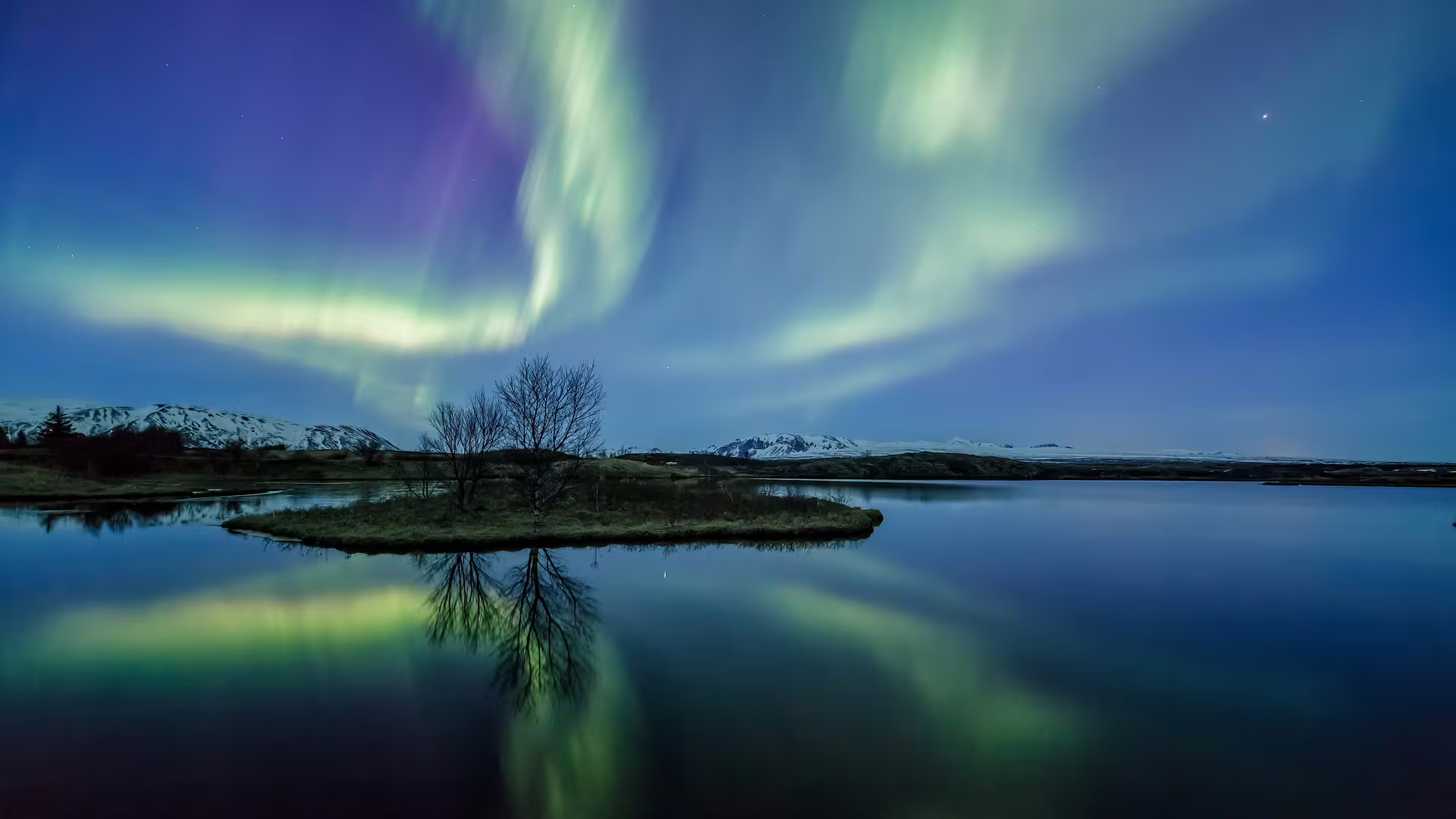 Aurora borealis dancing over a calm lake with small island and snowy mountains in Reykjavik, Iceland