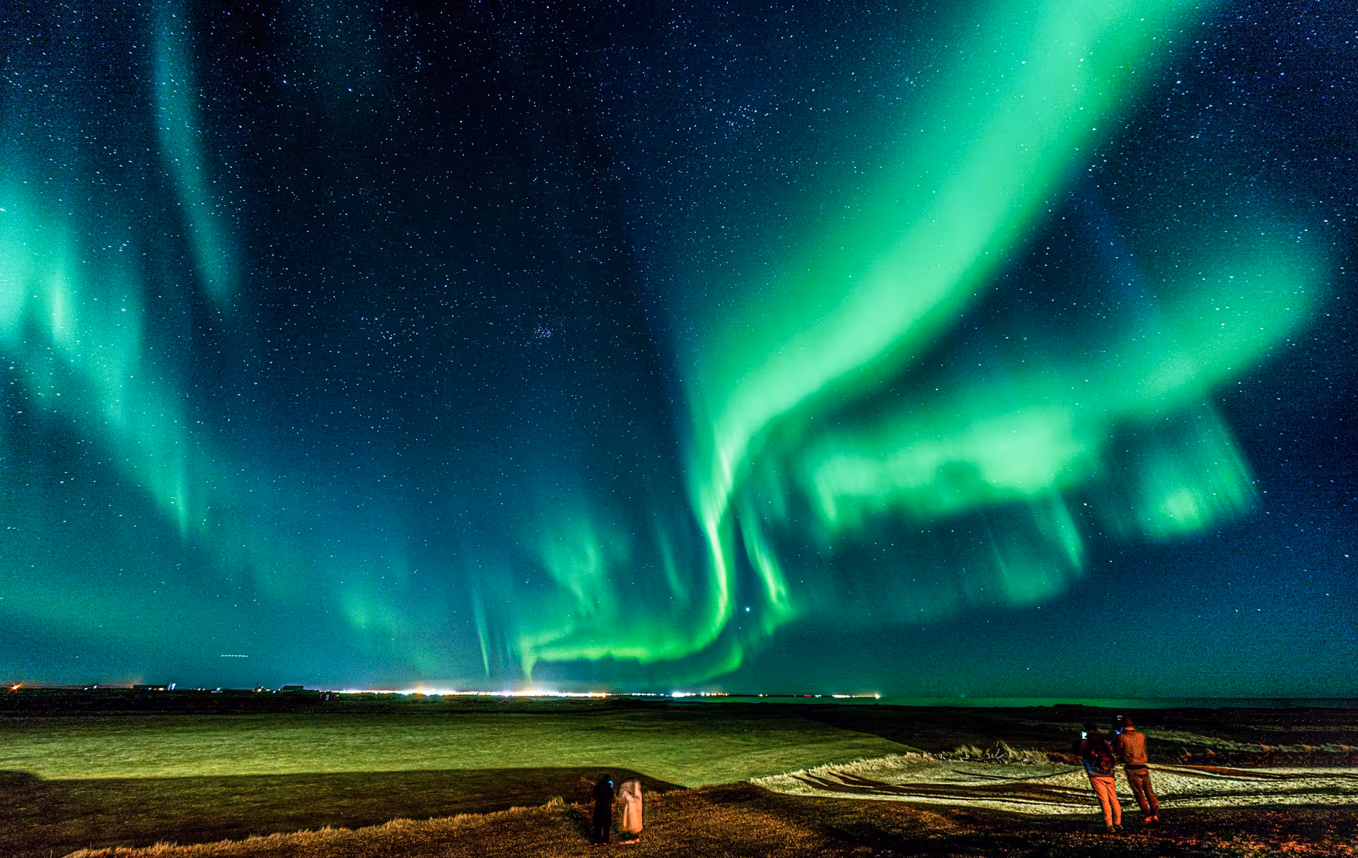 Vivid green northern lights aurora borealis over Iceland flatlands with two observers in foreground