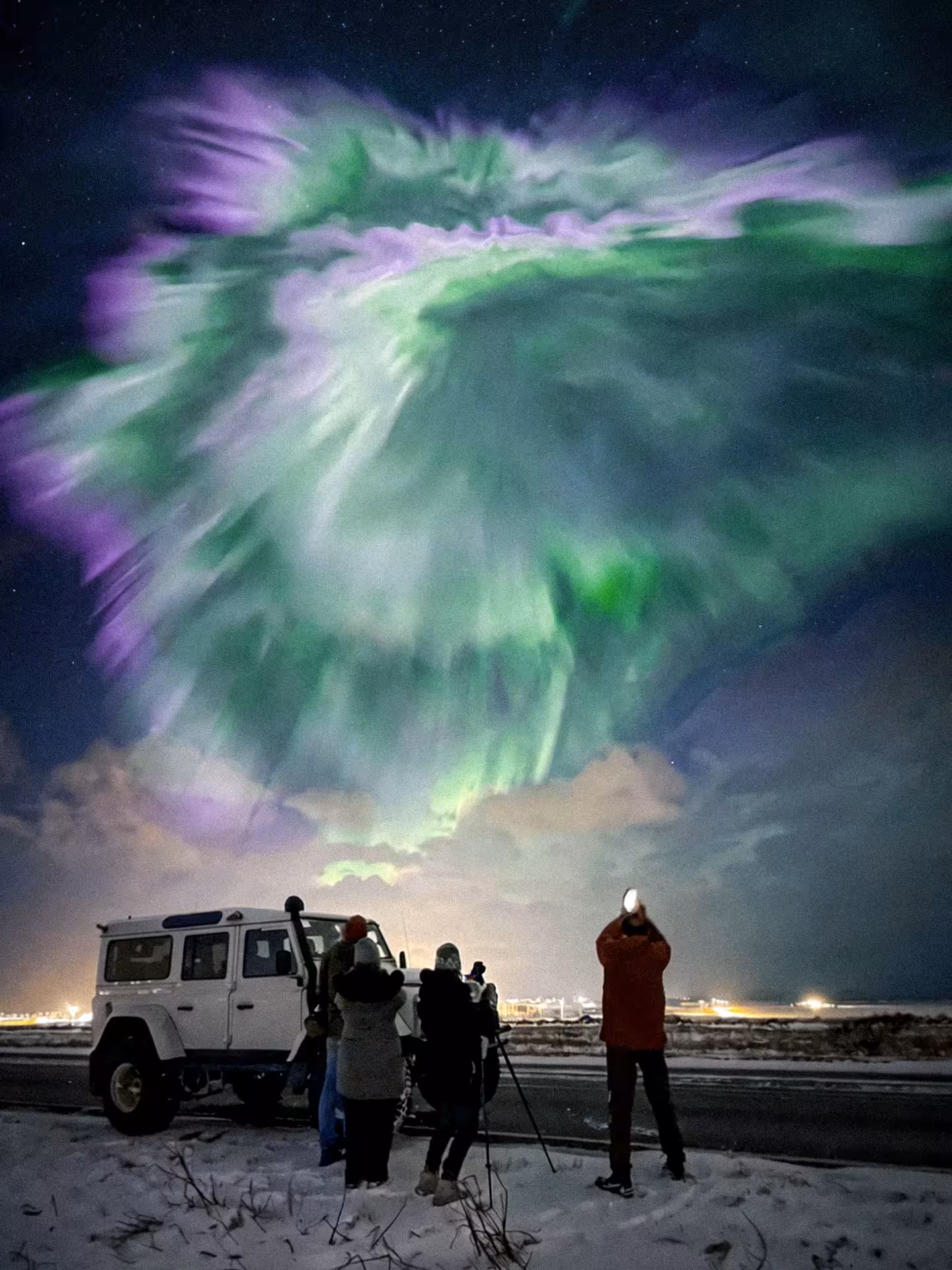 Group of tourists photographing vivid green and purple northern lights corona display over snowy Iceland landscape at night