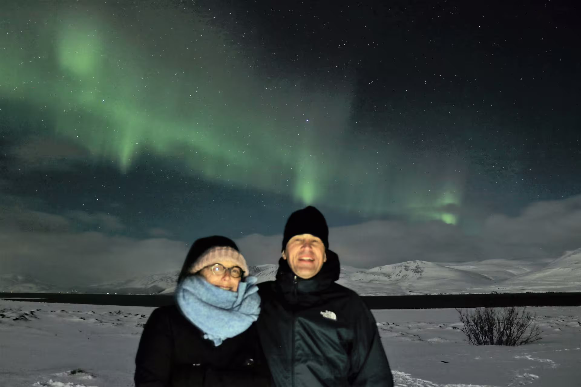 Couple posing under vivid green northern lights above snowy Icelandic landscape near Akureyri at night