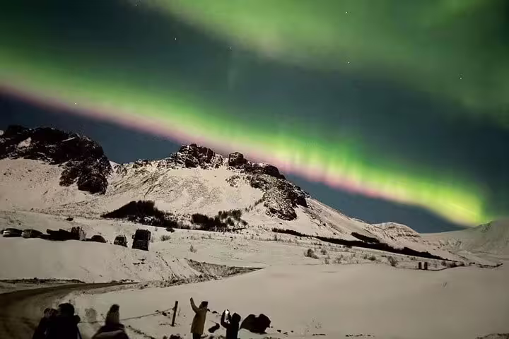 Tourists photographing vivid green aurora borealis dancing over snow-covered mountains at night