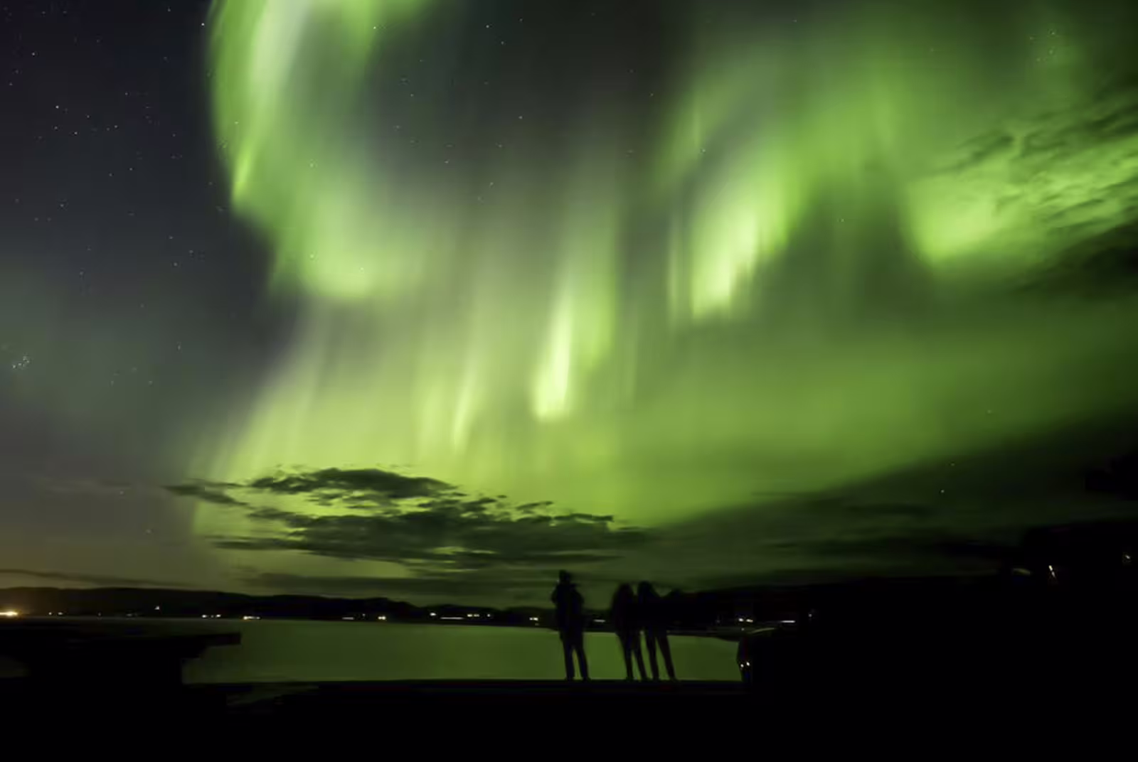 Three silhouetted people watching vibrant green northern lights aurora borealis over a calm lake in Iceland