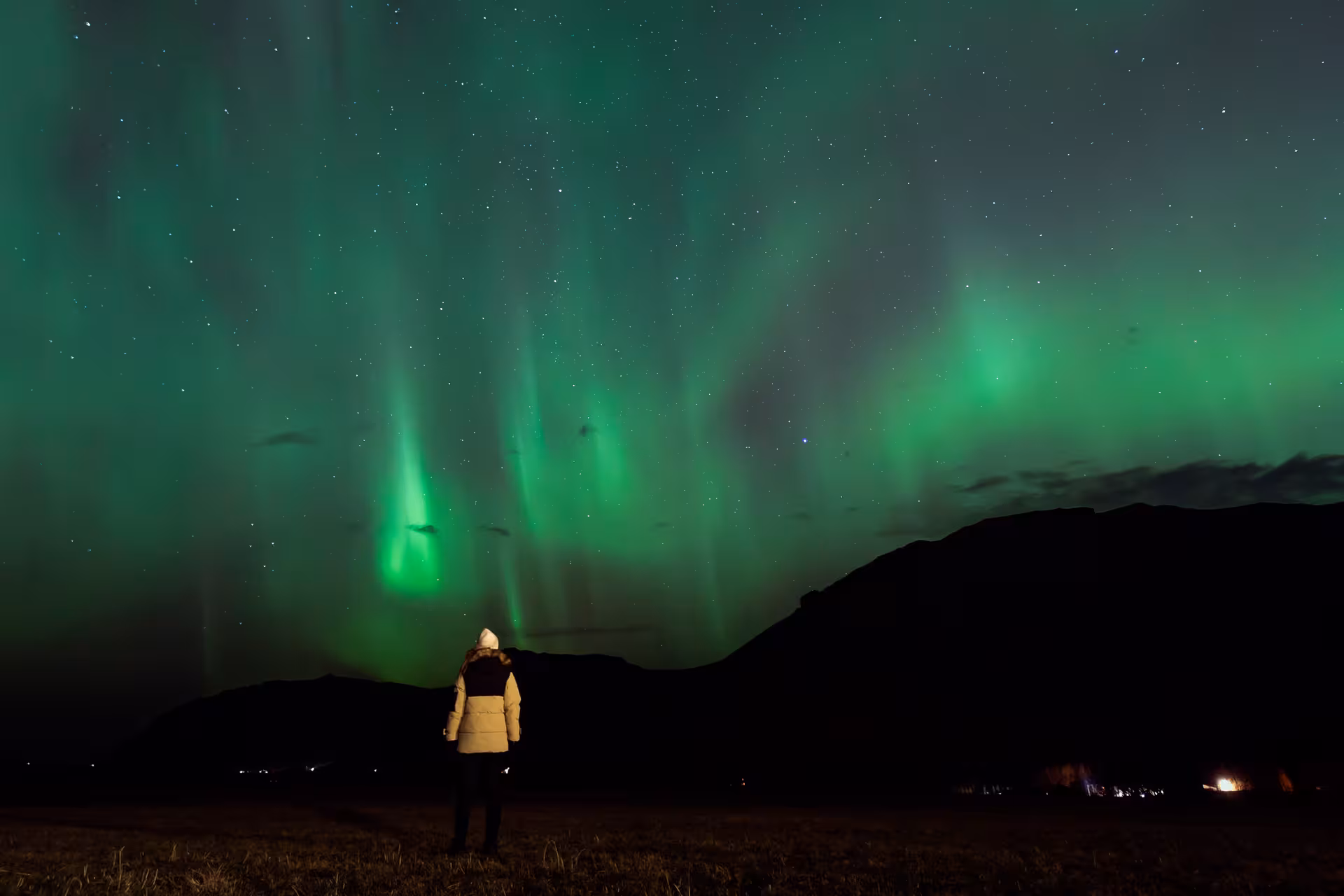 Person in winter jacket watching vivid green northern lights over Icelandic mountains at night