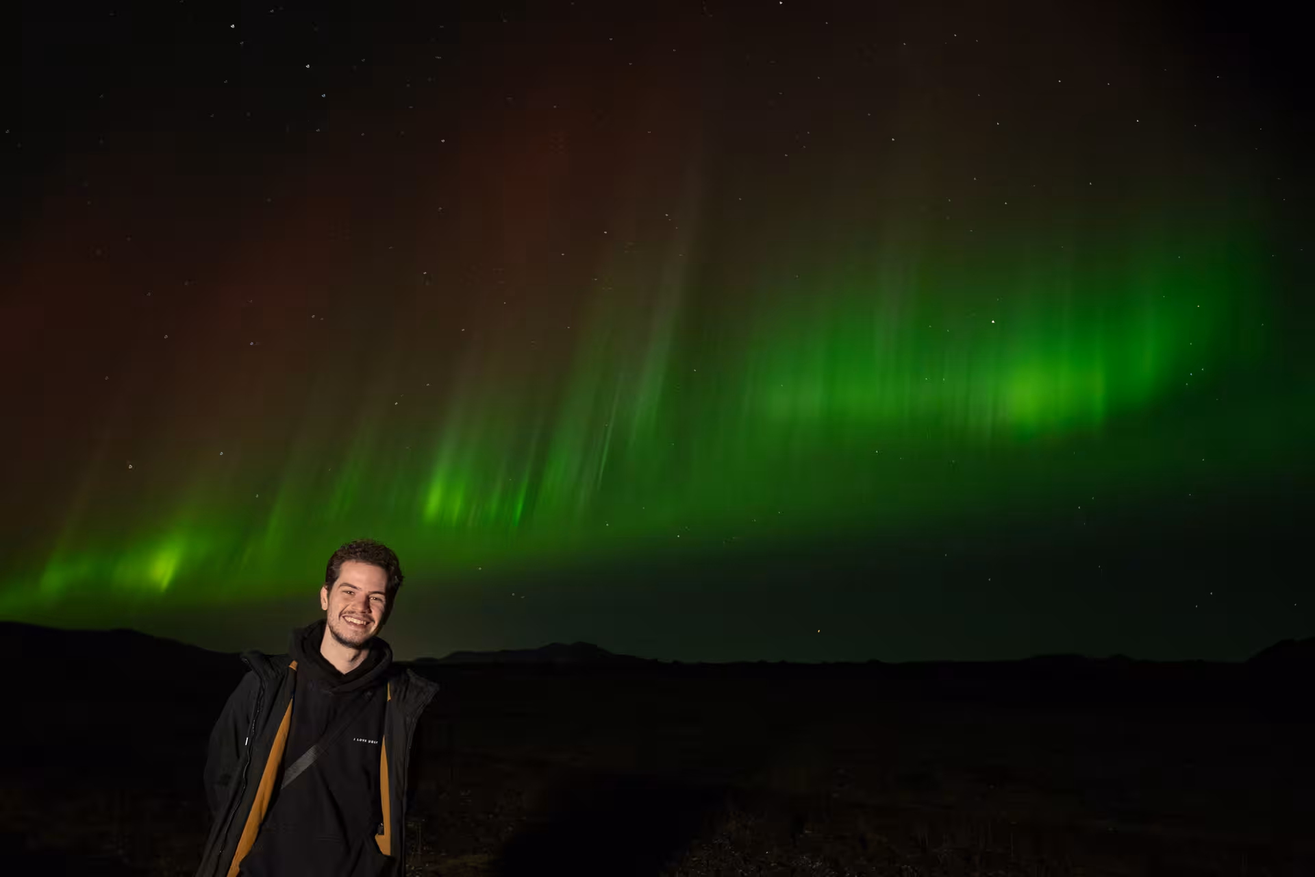 Smiling man posing in front of vibrant green and red northern lights over Iceland's dark landscape
