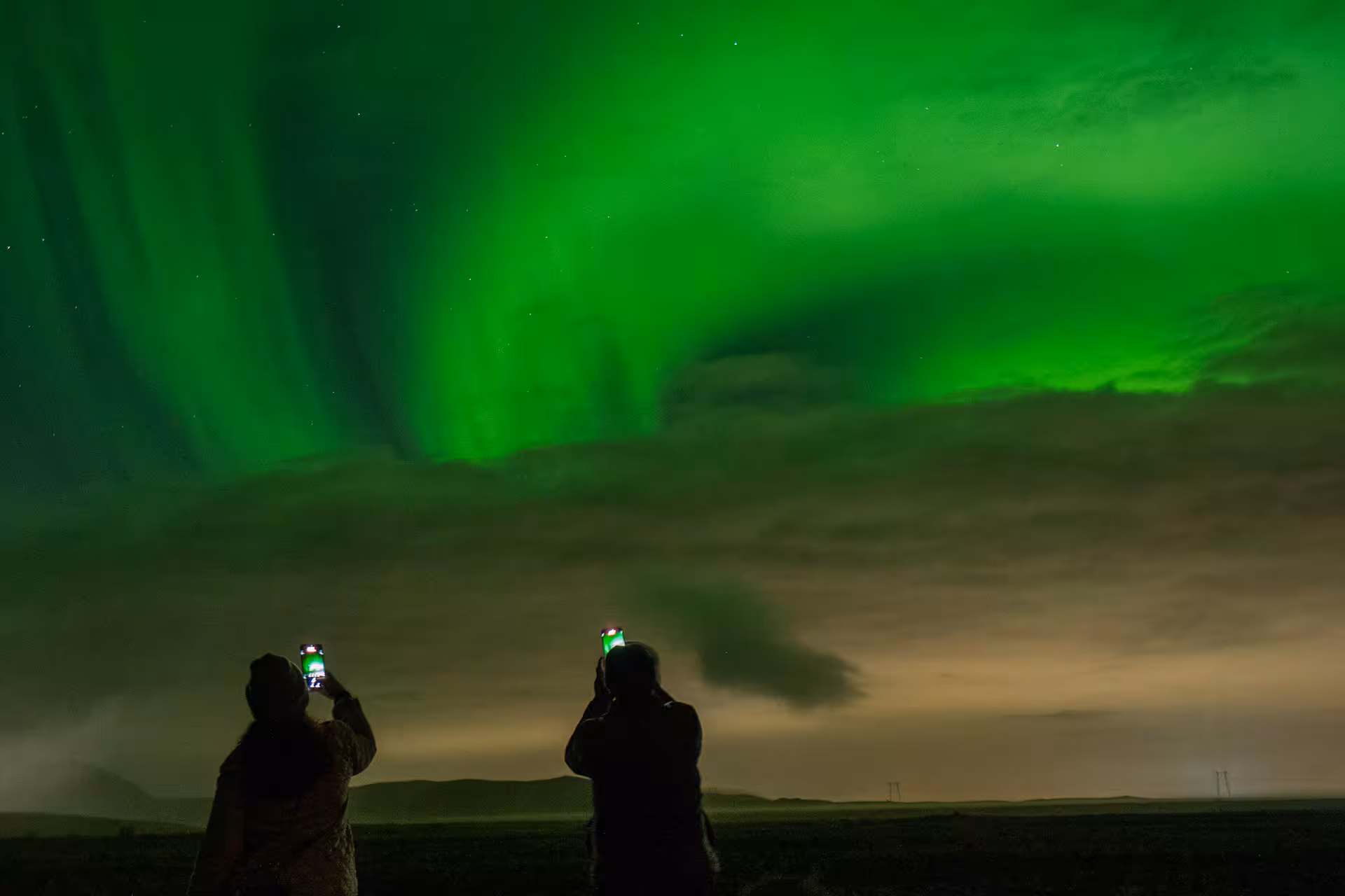Two tourists photographing vivid green northern lights with smartphones near Reykjavik at night