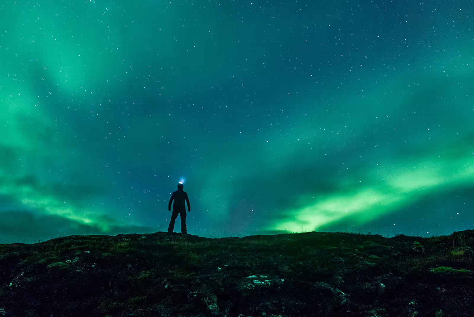Silhouetted person with headlamp standing on hill beneath vibrant green northern lights in Iceland night sky