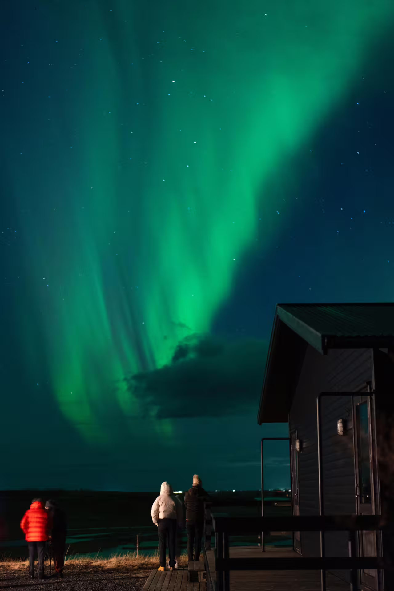 Three people watching vibrant green northern lights over Iceland from a wooden deck near a cabin at night