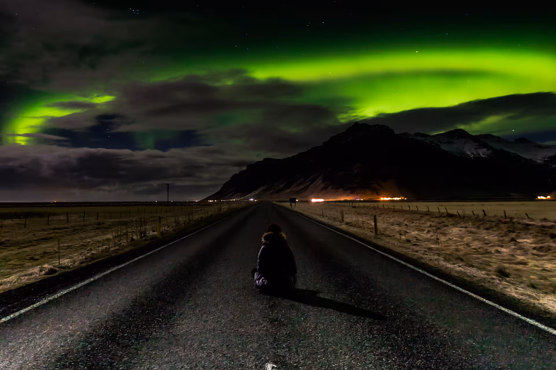 Person sitting on road watching vivid green northern lights over Icelandic mountains at night