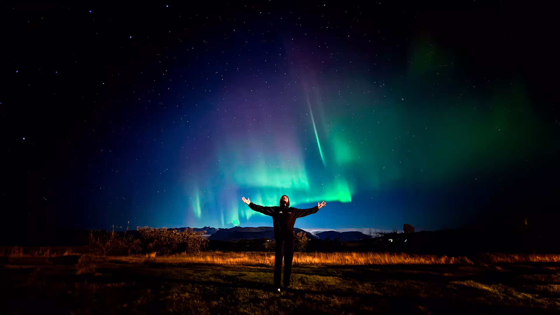 Person with arms outstretched under vibrant green and purple northern lights in Iceland night sky