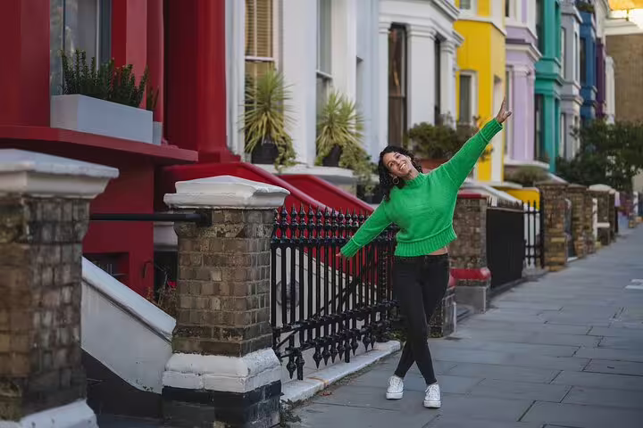 A joyful woman in a green sweater poses in front of vibrant, colorful houses on a charming street in Notting Hill, London.