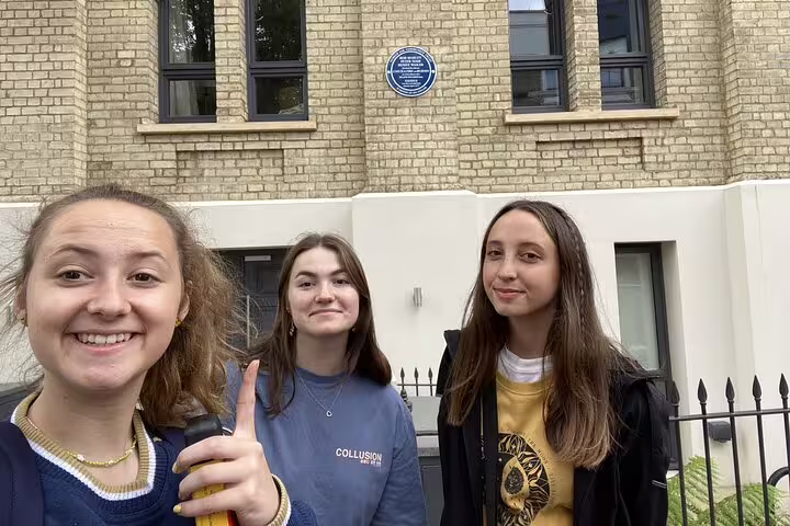 Three young women smiling in front of a blue heritage plaque on a brick building in Notting Hill