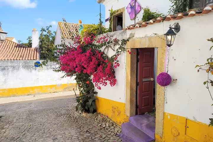 Charming Óbidos street with vibrant bougainvillea, colorful walls, and rustic cobblestone path under a sunny sky.