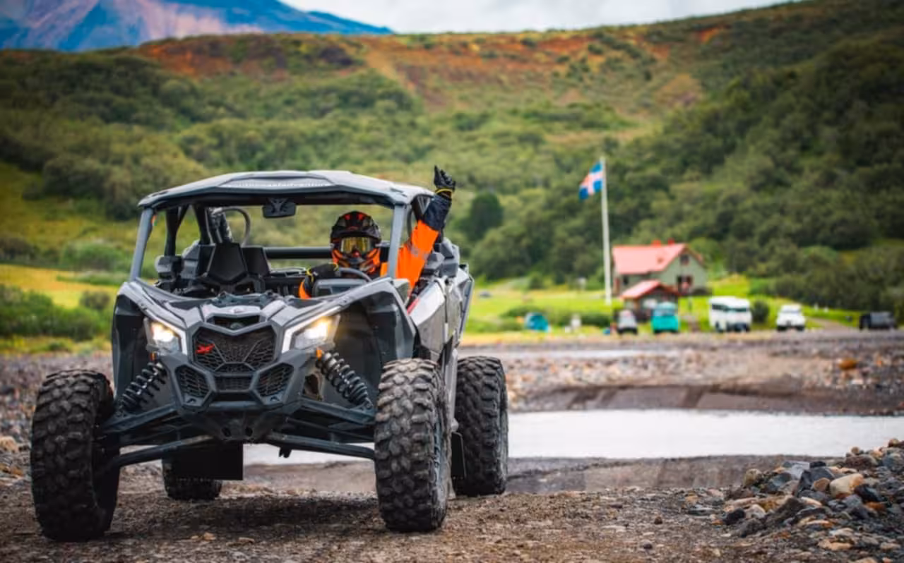 Driver in orange gear waving from a Can-Am buggy on rocky Iceland terrain with green hills behind