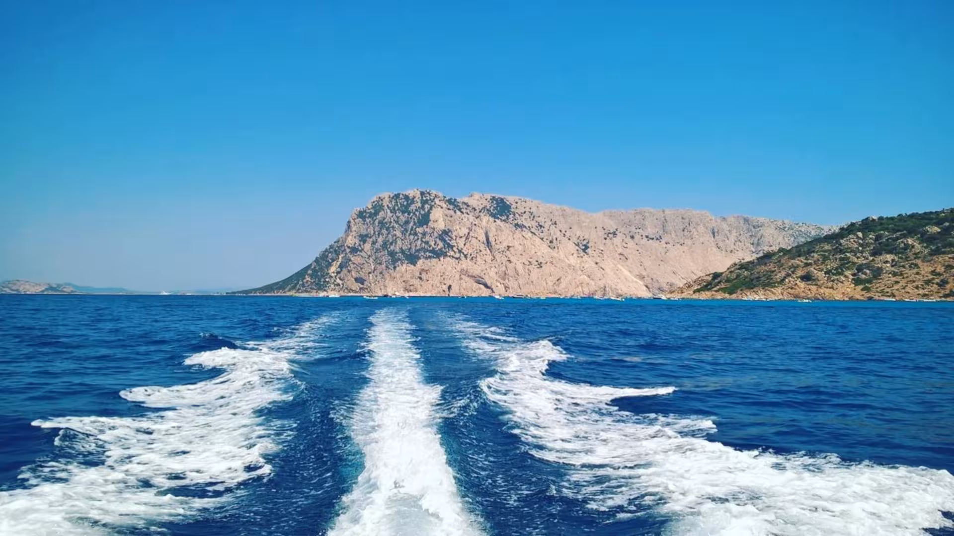 Boat wake trailing behind as it speeds towards Tavolara Island, with stunning rocky cliffs in the background.