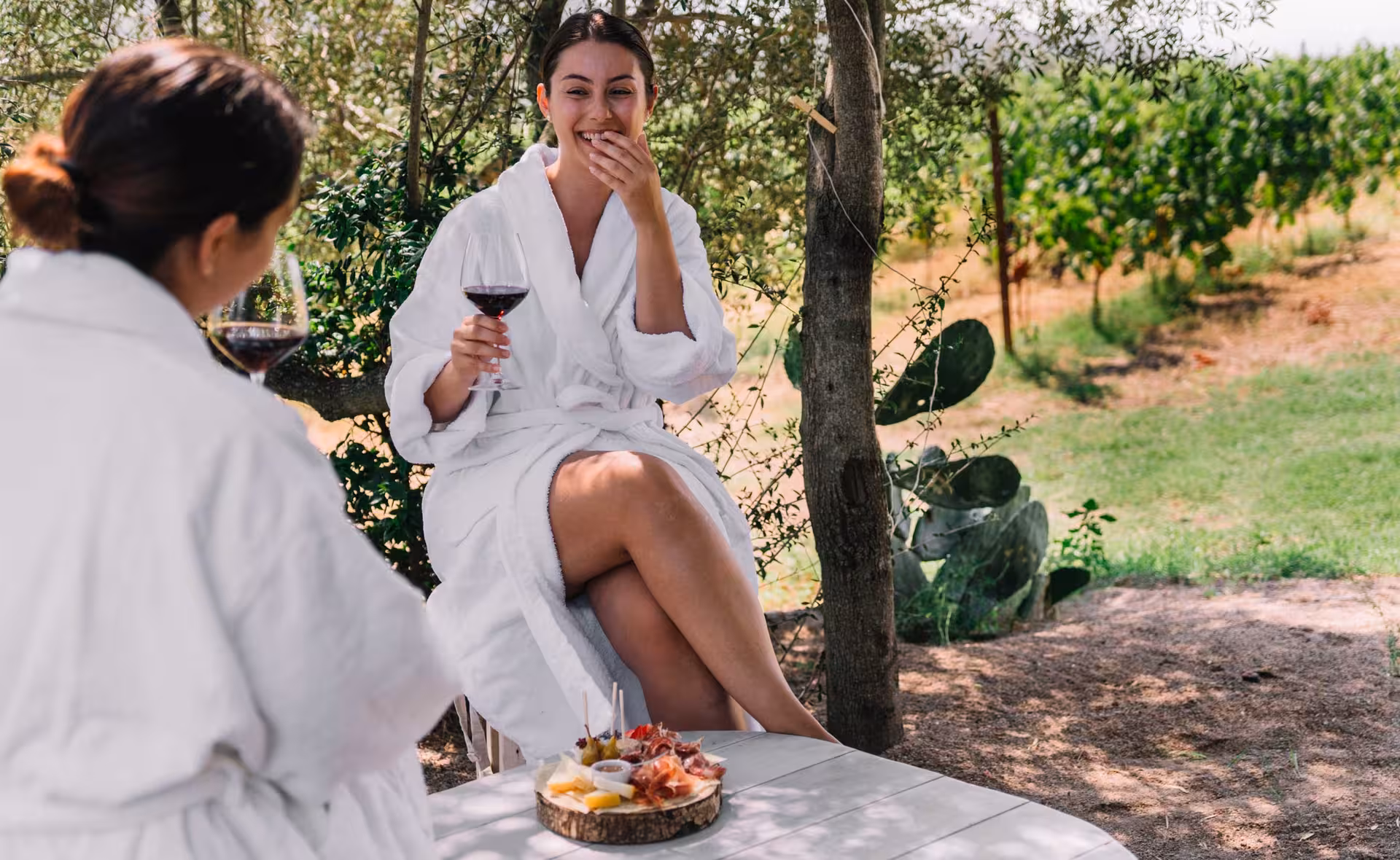 Women in white robes savoring red wine and snacks at a scenic vineyard table in a Gallura winery, Olbia.