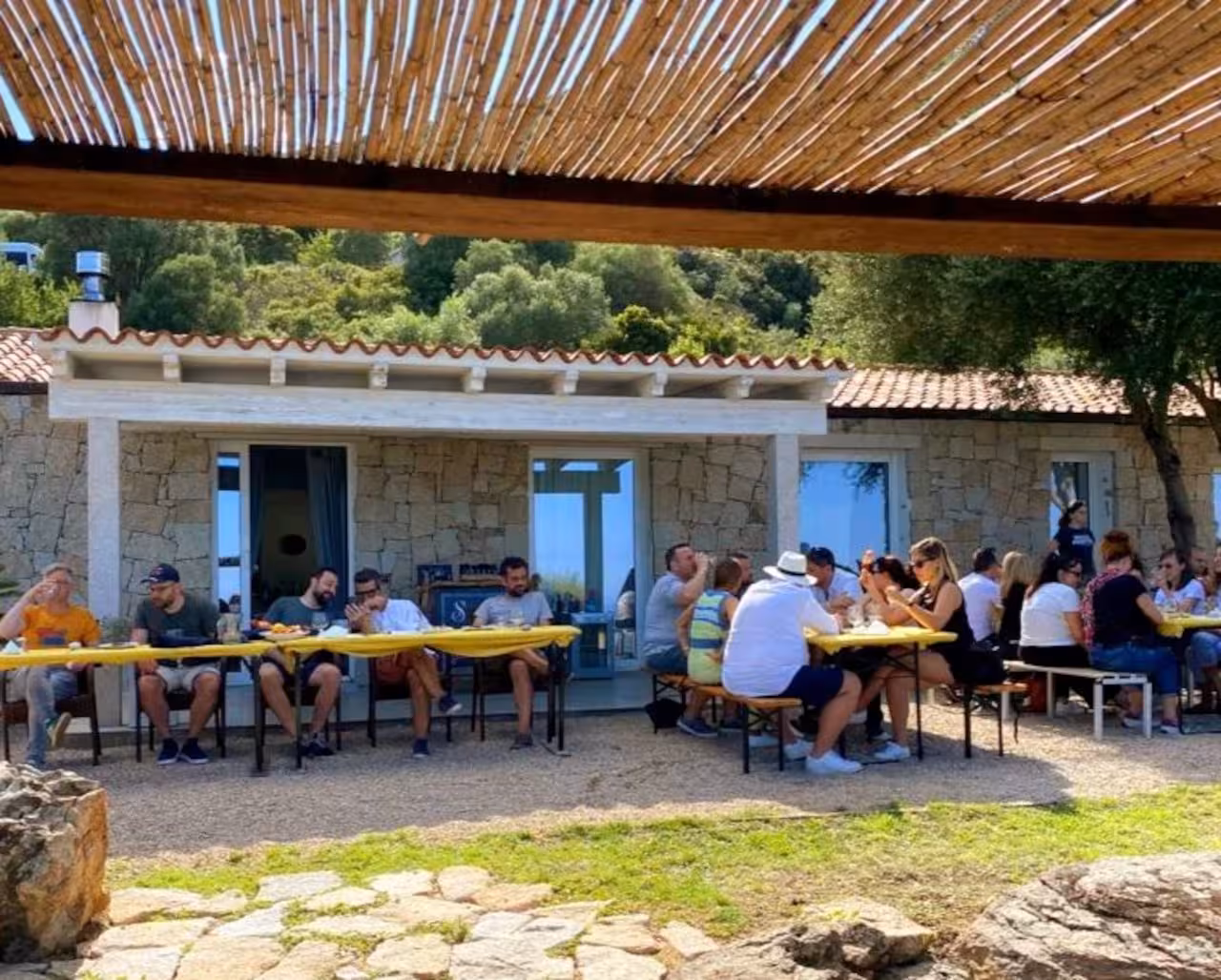 Visitors dining in a rustic setting at a myrtle-making factory in Olbia, surrounded by nature.