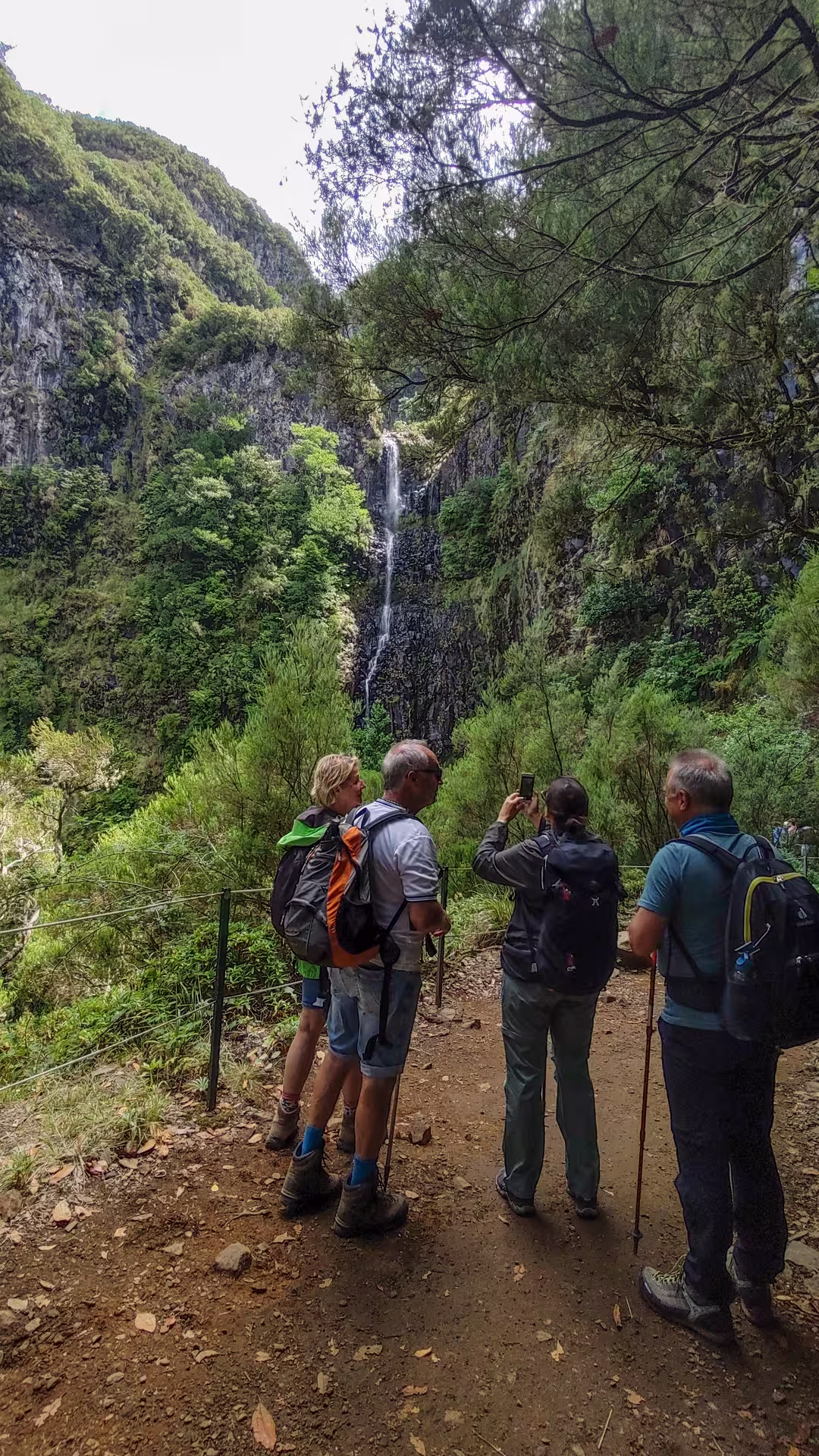 Hikers capturing the stunning waterfall view on The Old Levada’s Hike, amidst dense greenery and rocky cliffs.