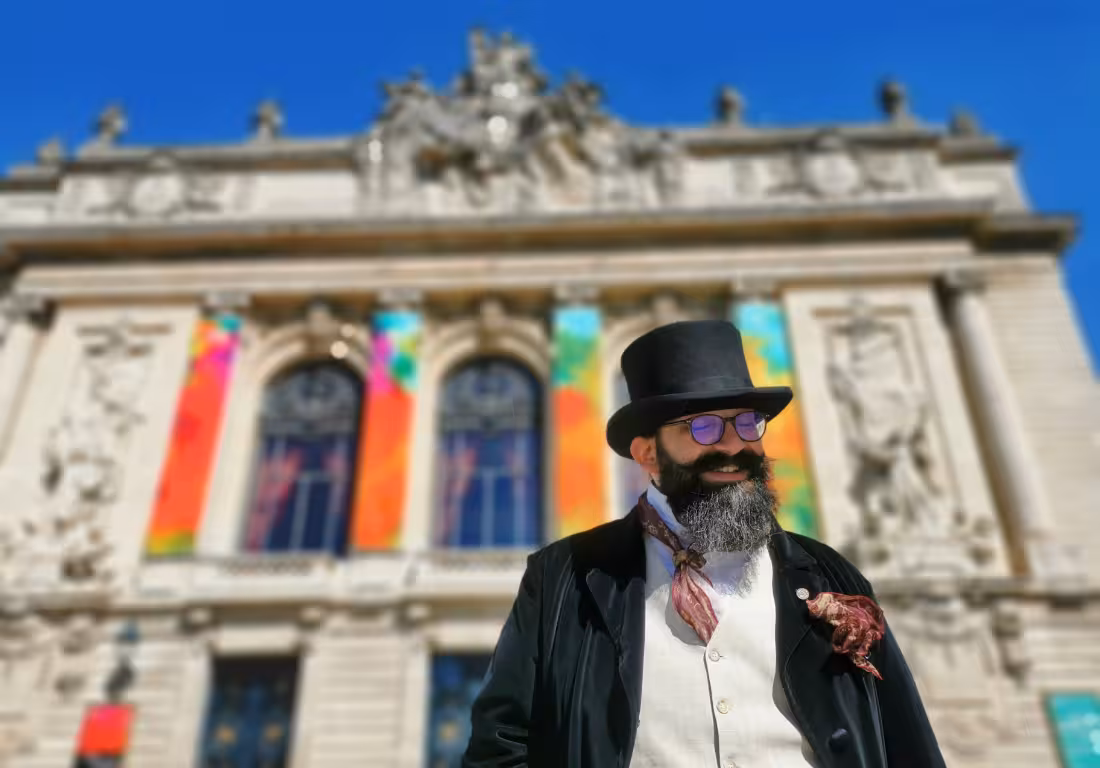 Bearded guide in Victorian top hat and waistcoat posing before ornate Lille building with colorful windows