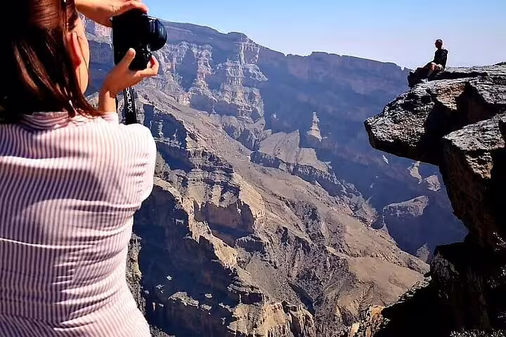 Tourist photographs a person sitting on a dramatic cliff edge at Oman's breathtaking Grand Canyon.