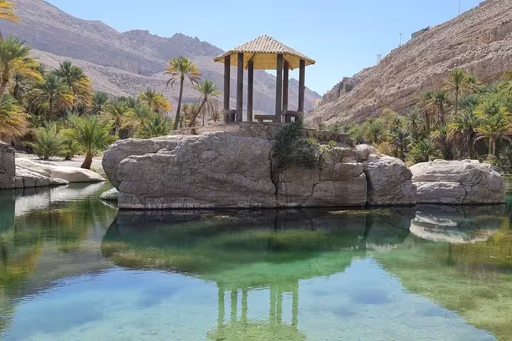 Scenic view of a gazebo on a rock surrounded by palm trees and crystal-clear waters in an Omani wadi.