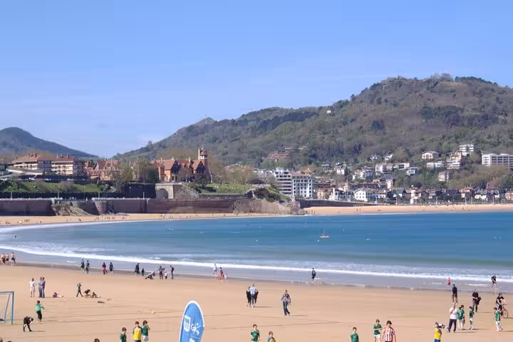 People enjoy a sunny day at the scenic Ondarreta Beach in San Sebastián, with lush hills in the background.