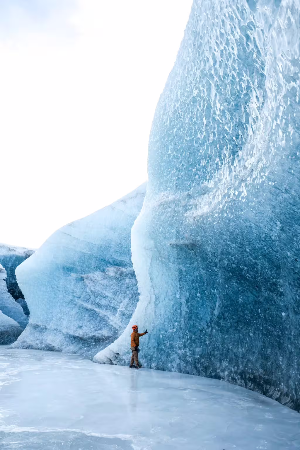 Climber in orange gear touches towering textured blue glacier wall during private ice cave climbing photoshoot adventure