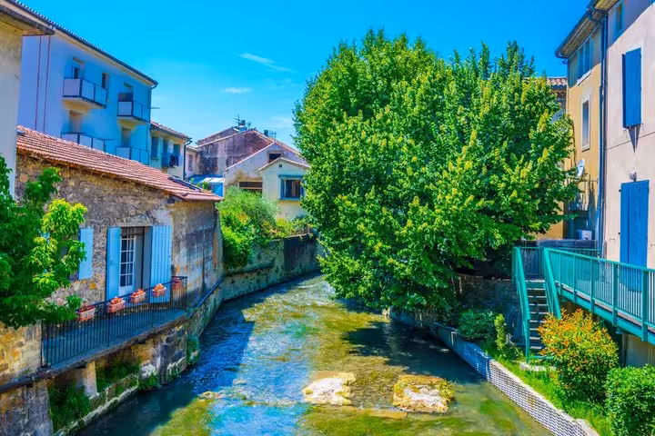 Narrow river canal flowing between old stone buildings and lush green trees in Orange, France
