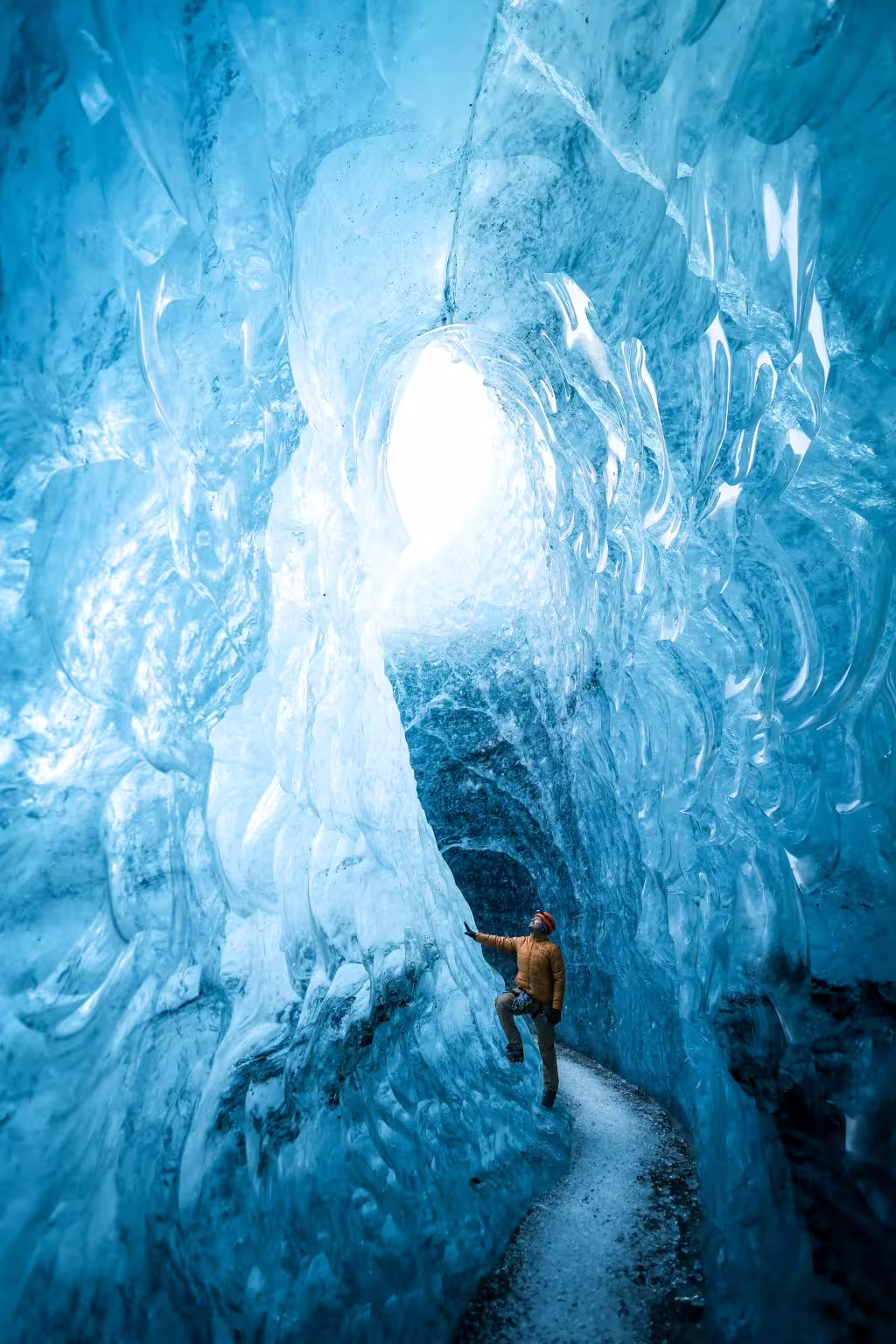 Explorer in orange helmet scaling glowing blue ice cave walls during a private glacier climbing photoshoot adventure
