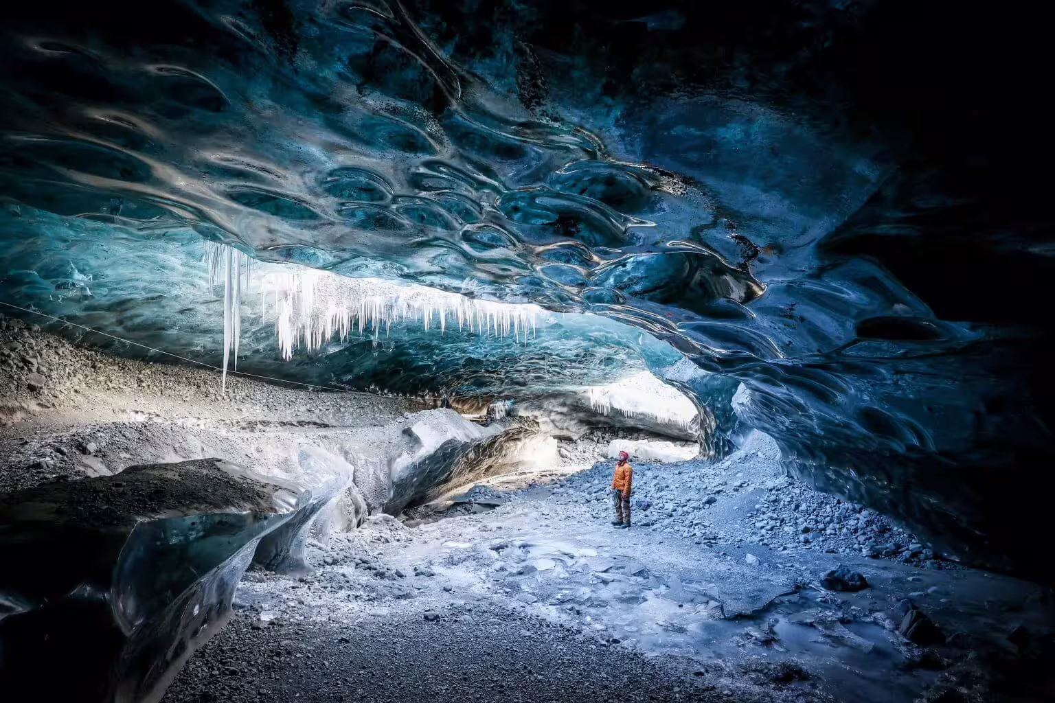Explorer in orange jacket marveling at massive glacier ice cave with icicles on a private photography tour