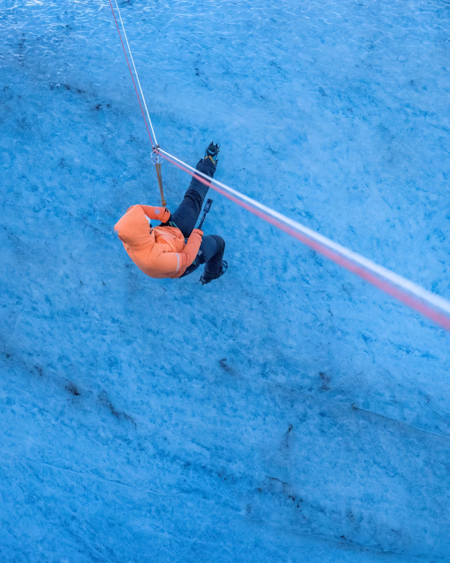 Aerial view of adventurer in orange jacket zip lining over stunning blue glacier surface in Iceland winter tour