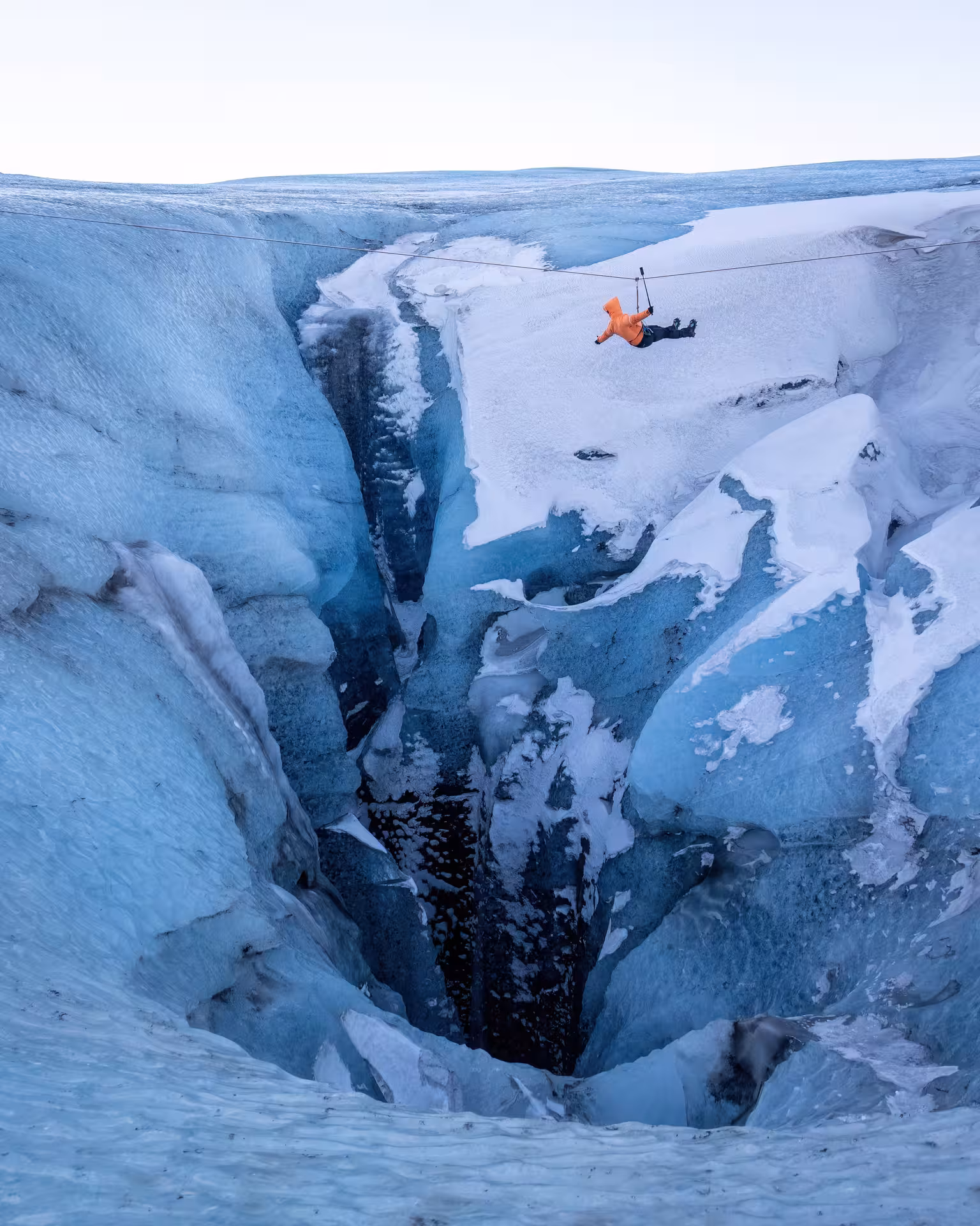 Adventurer in orange jacket zip lining over a deep glacier crevasse on a winter Iceland glacier tour
