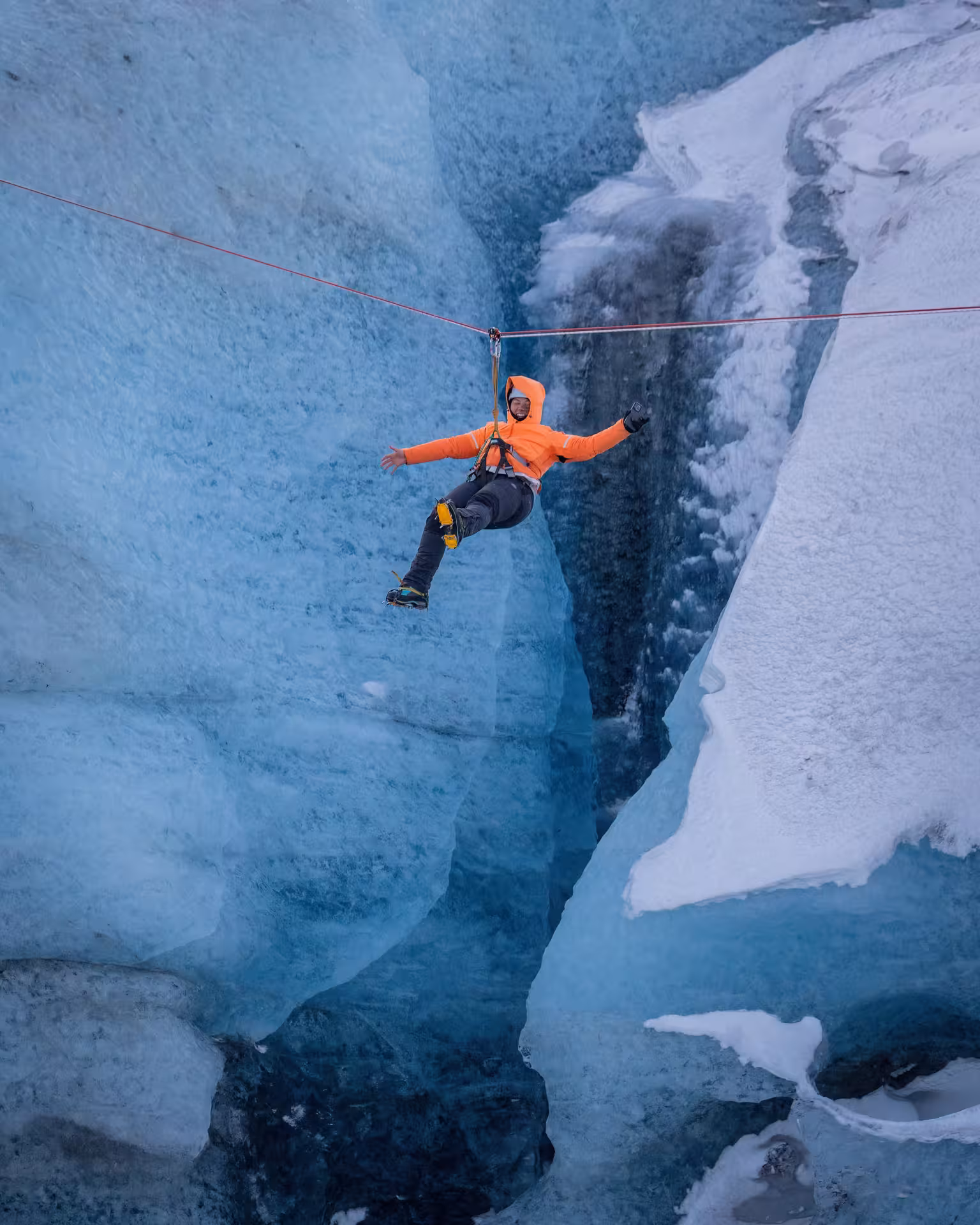 Adventurer in orange jacket zip lining over a deep glacier crevasse on an Iceland winter tour