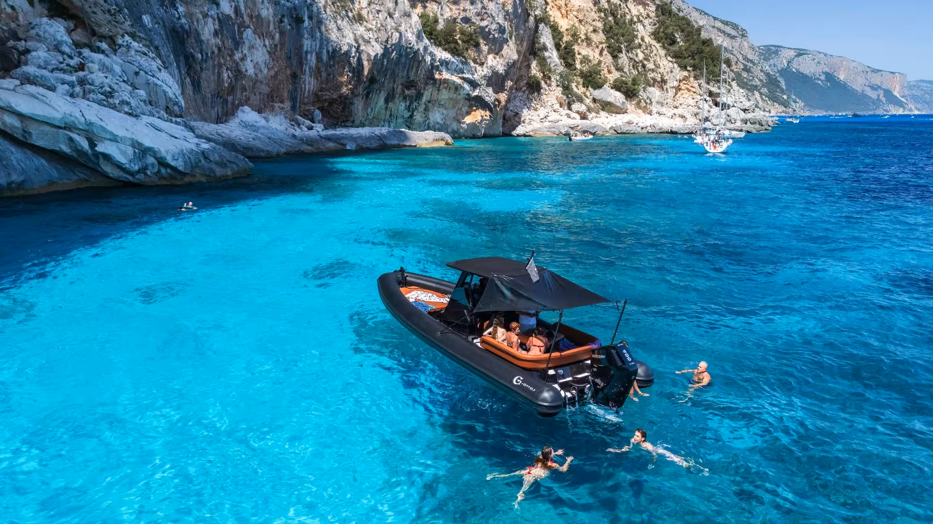 Visitors swim near a dinghy in the pristine waters of Cala Goloritzè, surrounded by dramatic rocky cliffs.